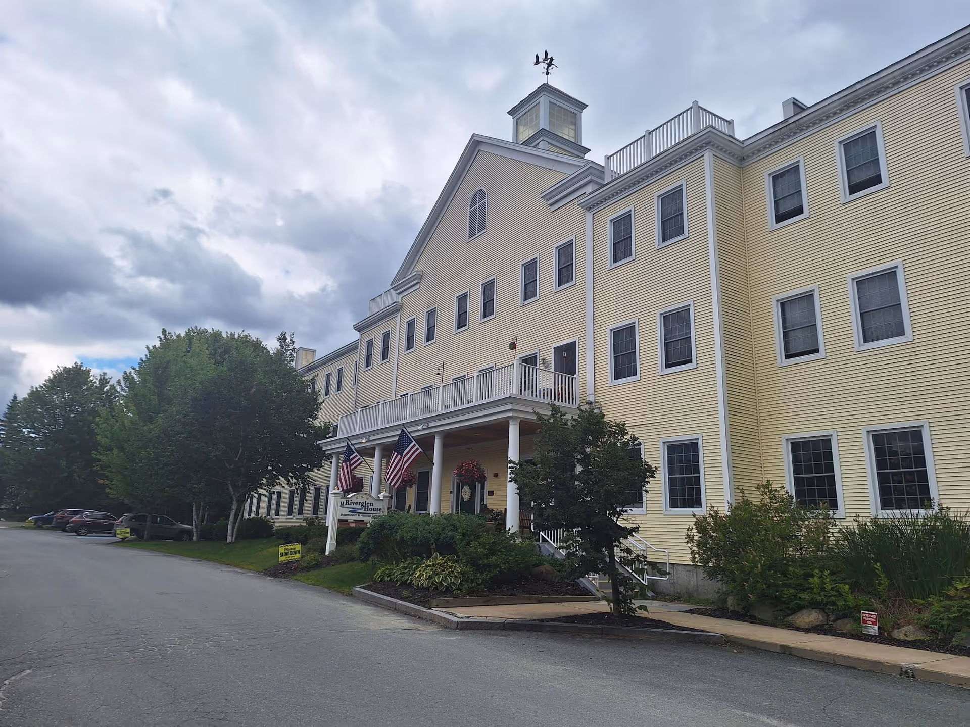 Exterior view of a large, three-story yellow building with white trim and multiple windows. The building has a covered entrance with white columns, American flags, and a sign that reads 'Riverglen House'. There are trees and shrubs around the entrance and a parking area with several cars on the left side. The sky is cloudy.