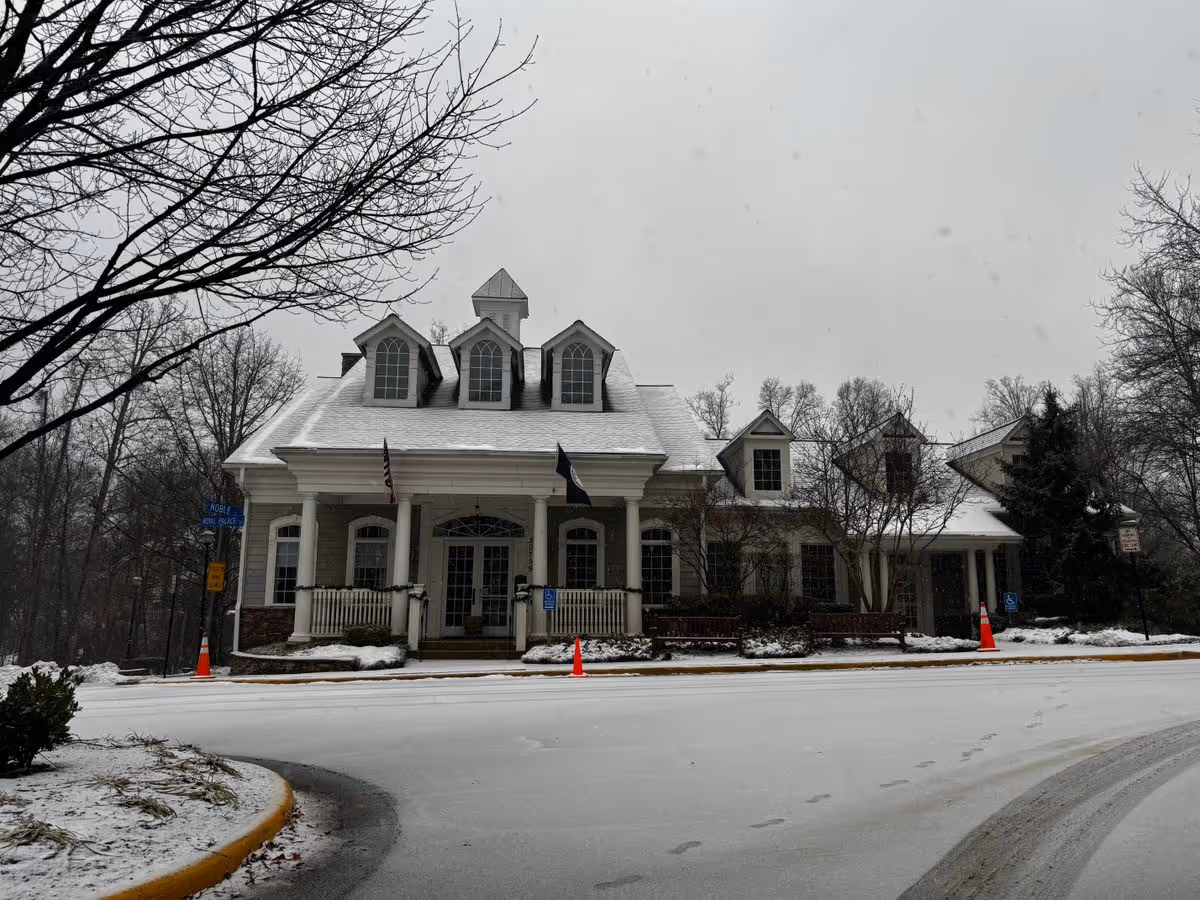 Snow-covered front entrance of a large colonial-style building with dormer windows, flags, and orange traffic cones.