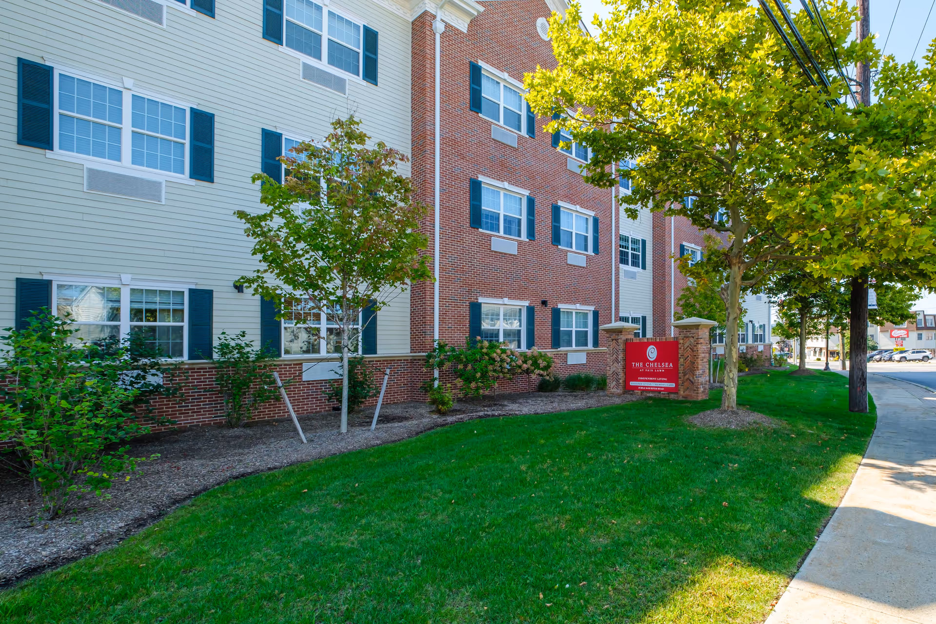 Exterior front of a brick-and-siding senior living building with landscaping and a red sign for The Chelsea at Fair Lawn.