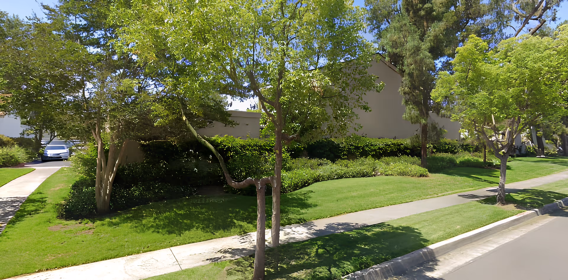 Well-maintained grassy lawn and sidewalk lined with trees and shrubs in front of a low beige building, with a white car parked at the left.
