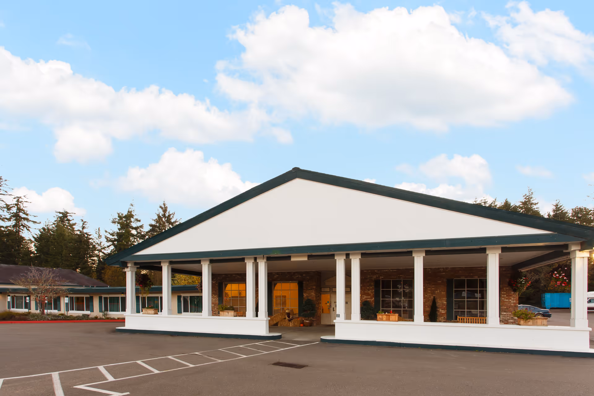 Front exterior view of Life Care Center of Coos Bay showing a single-story building with a large covered entrance supported by white columns, brick walls, and windows. The sky is partly cloudy and there are trees in the background.