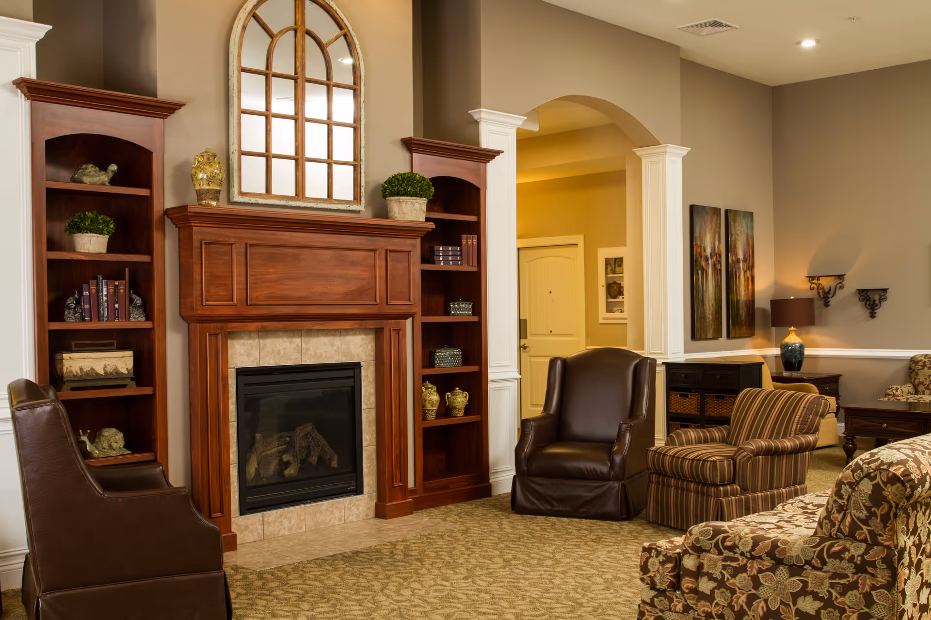 A cozy senior living facility common area featuring a wooden fireplace with built-in shelves on either side, decorated with books, plants, and ornaments. The room has comfortable seating including leather and striped armchairs, floral patterned sofas, warm beige walls, and soft lighting from wall sconces and a table lamp.