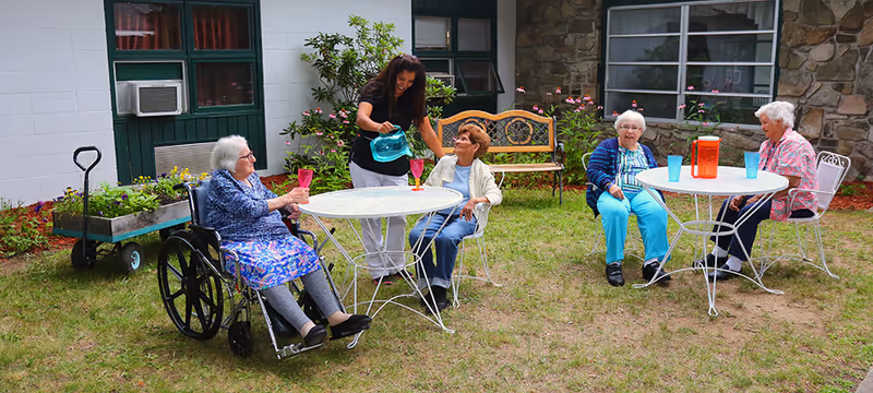 A caregiver pours drinks for four elderly women seated at white metal patio tables in a grassy courtyard outside a senior center.