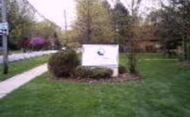 Lawn and sidewalk with a white sign surrounded by bushes in front of a facility
