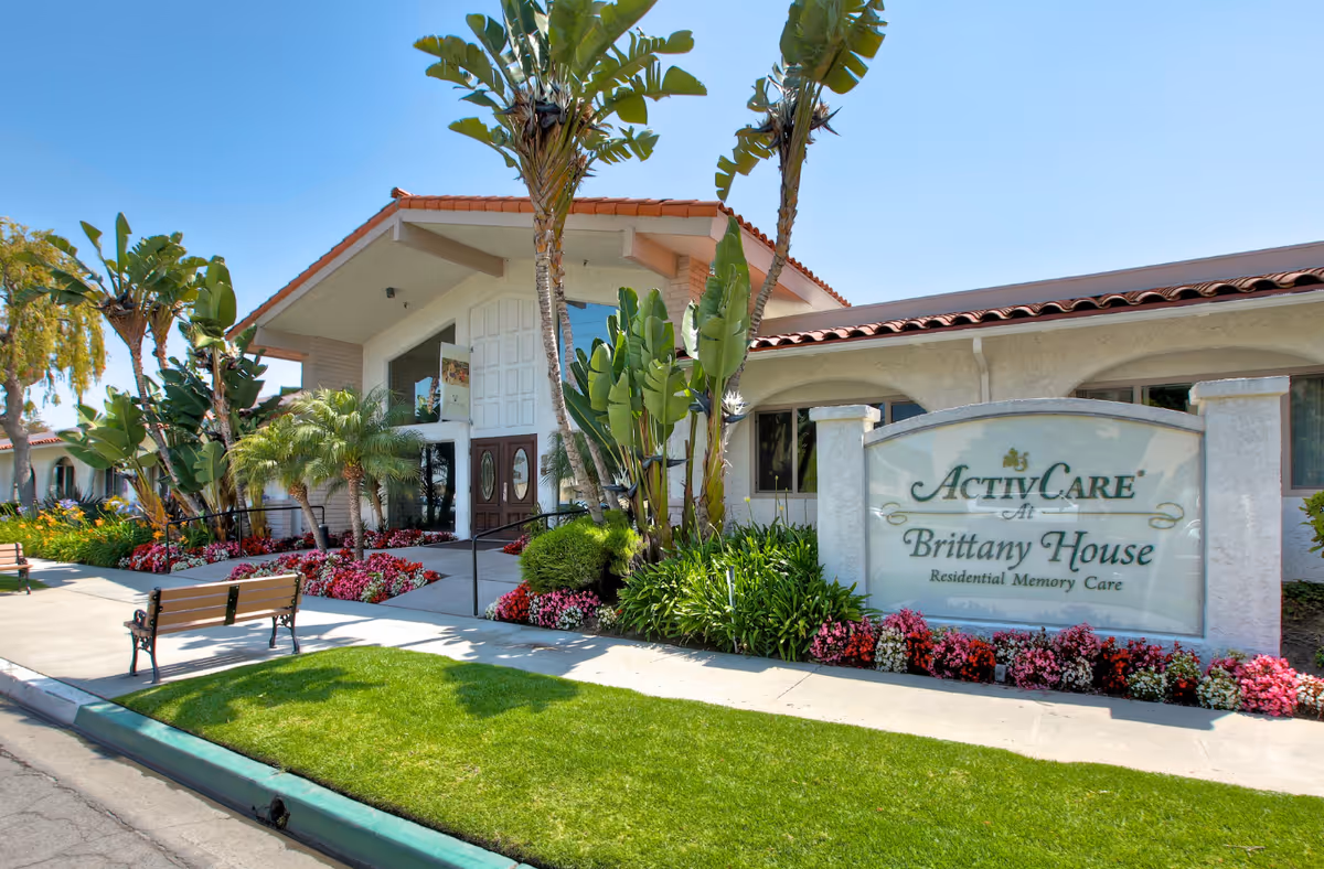 Exterior view of Brittany House Residential Memory Care facility with a well-maintained garden, palm trees, colorful flowers, benches, and a large sign displaying the facility's name. The building has a tiled roof and large entrance doors under a peaked roof.