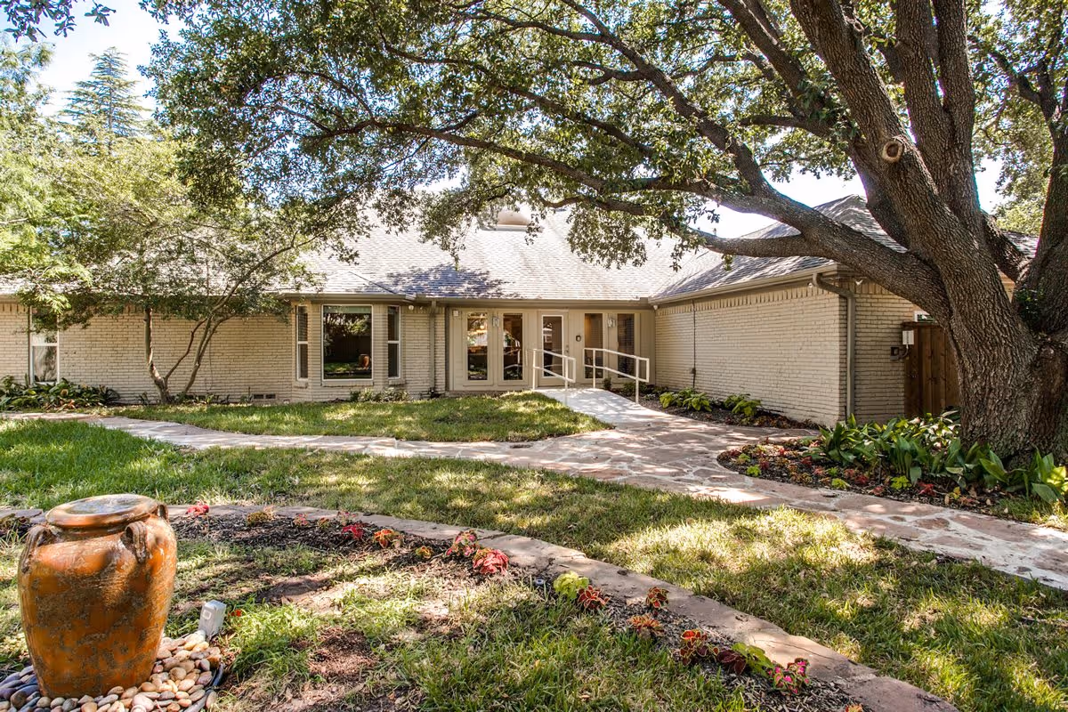 Outdoor view of a single-story brick building with a light-colored exterior, surrounded by a well-maintained garden with green grass, flower beds, and a large tree providing shade. A stone pathway leads to a ramp and double glass doors entrance.