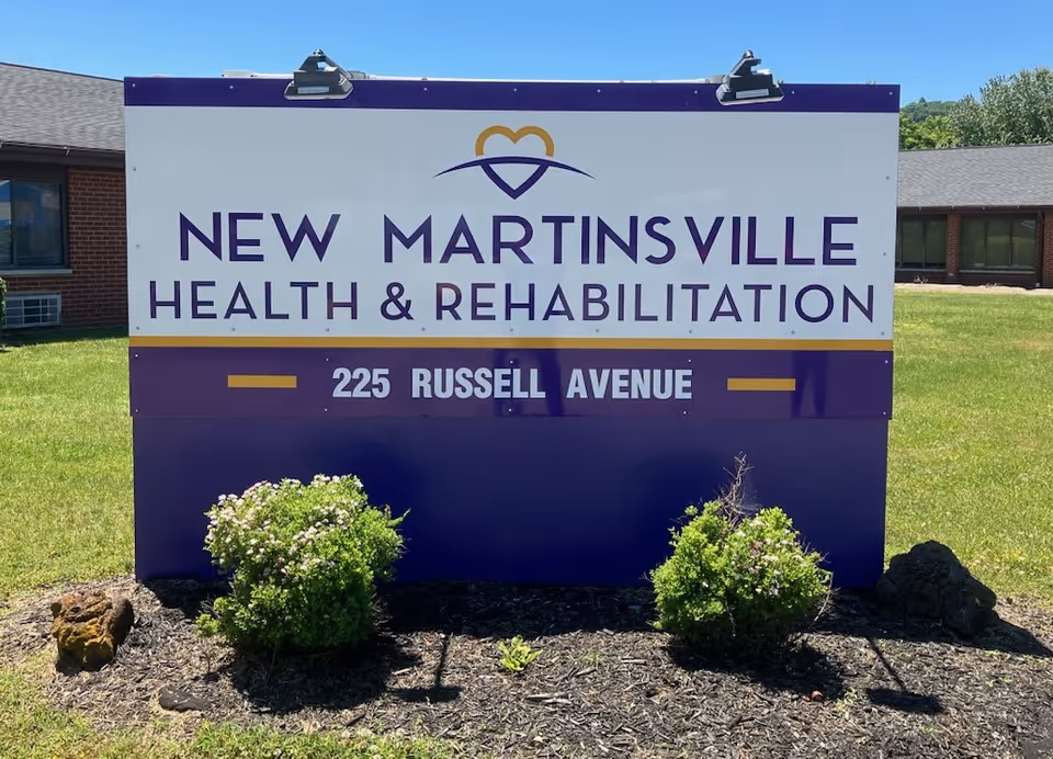 Outdoor sign for New Martinsville Health & Rehabilitation located at 225 Russell Avenue, with a building and green lawn in the background under a clear blue sky.