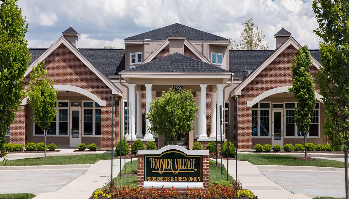 Front exterior view of Hoosier Village building with a brick facade, white columns at the entrance, and a sign in front that reads 'Hoosier Village Roosevelts & Green Onion'. The area is landscaped with small trees, bushes, and flowers under a partly cloudy sky.