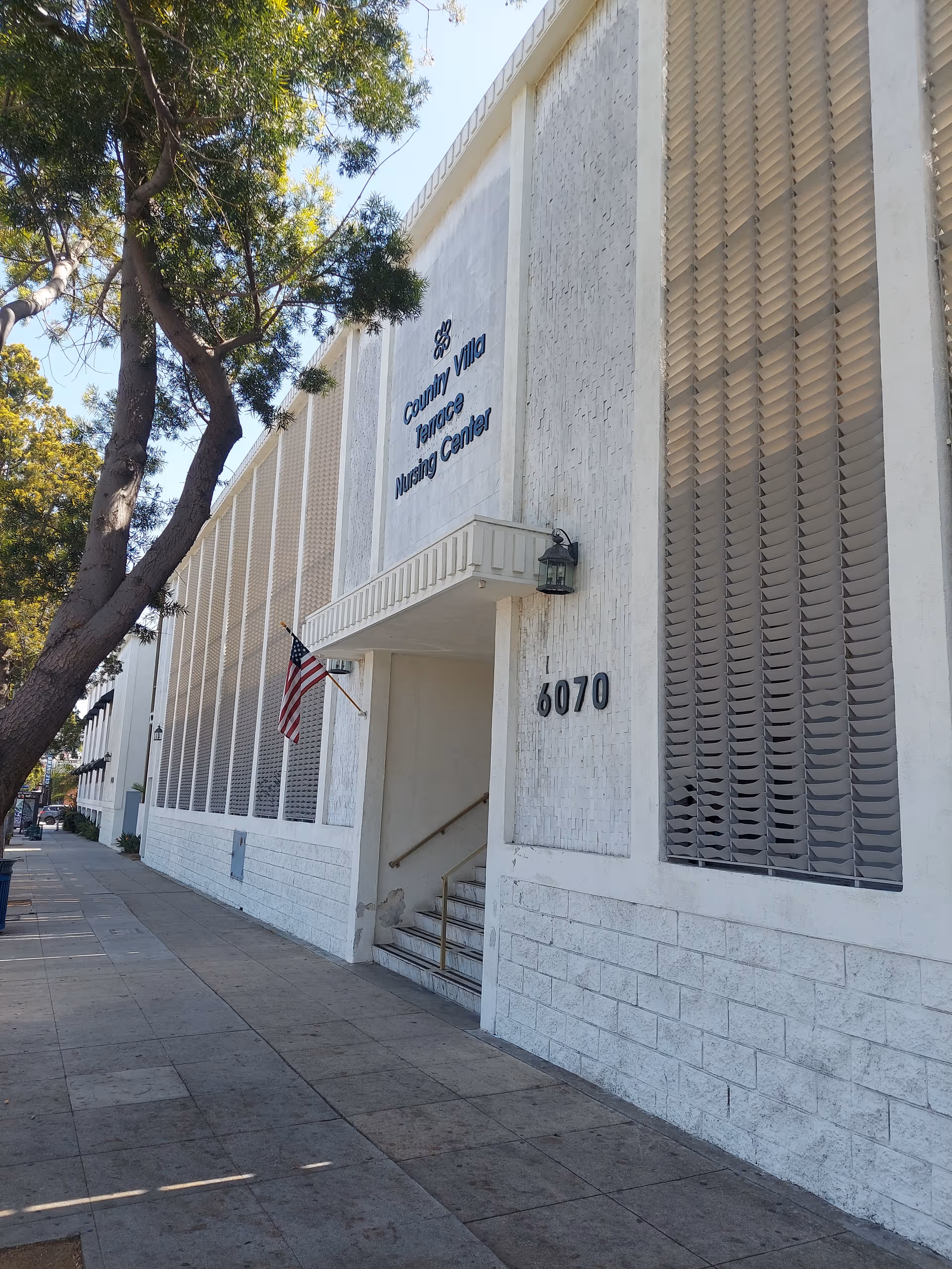 Sunlit exterior of a senior living center with stairs, flag, and 6070 address.