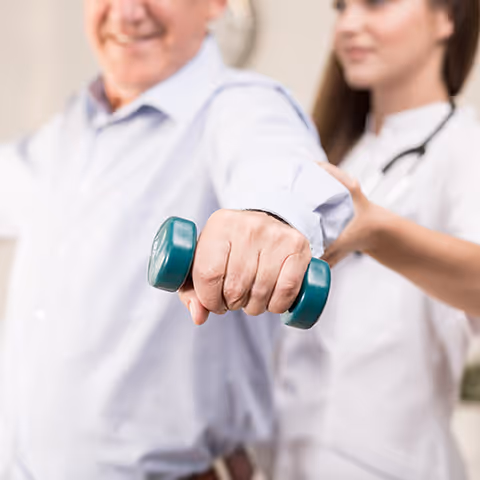 Caregiver assists an elderly resident as they lift a teal dumbbell in an indoor facility.