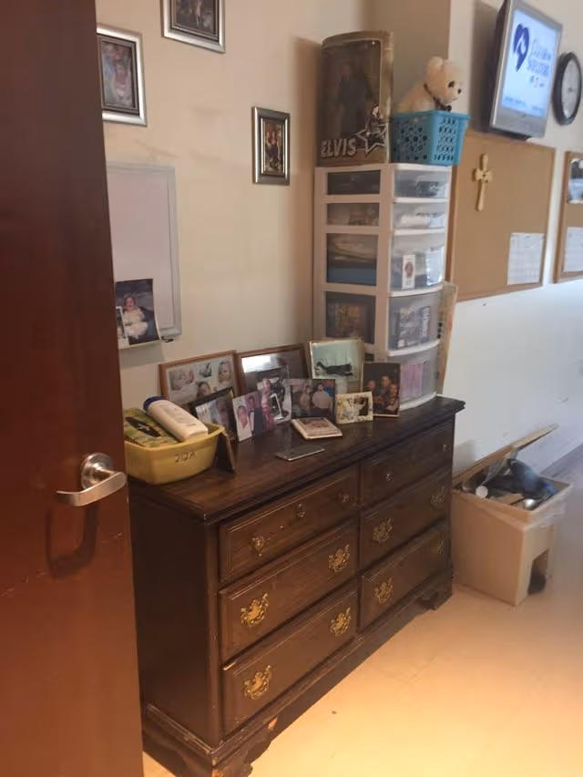 An interior room with a dark dresser, framed photos, and storage containers against the wall.