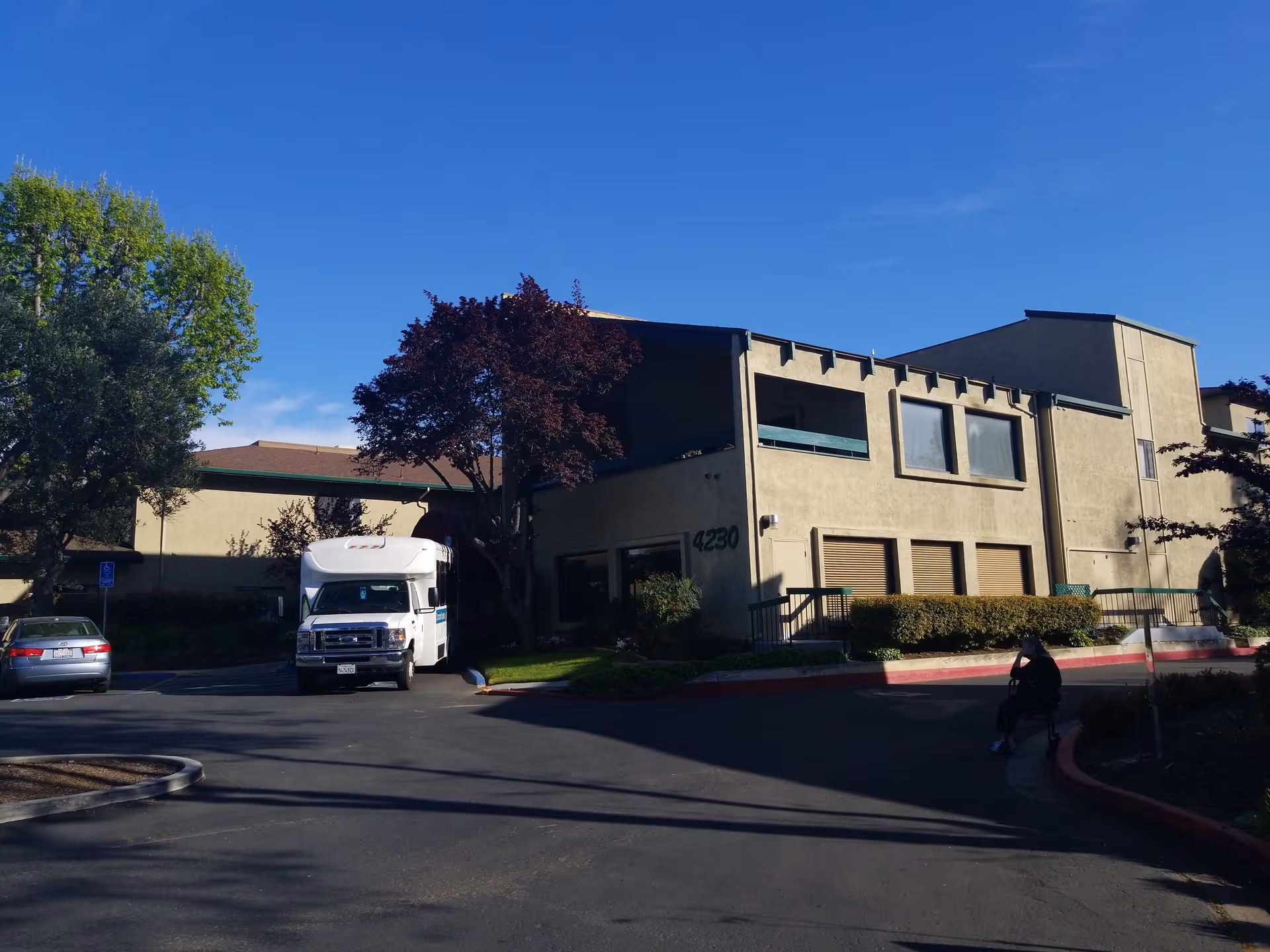 Front exterior of Concord Royale senior living with driveway, white shuttle bus, and a wheelchair user.