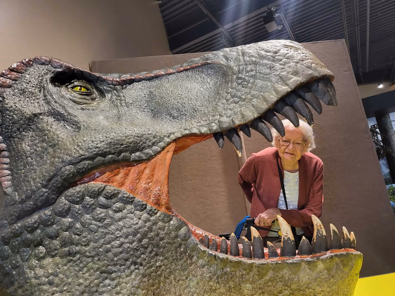 Elderly woman beside a giant dinosaur head sculpture indoors at a senior facility.