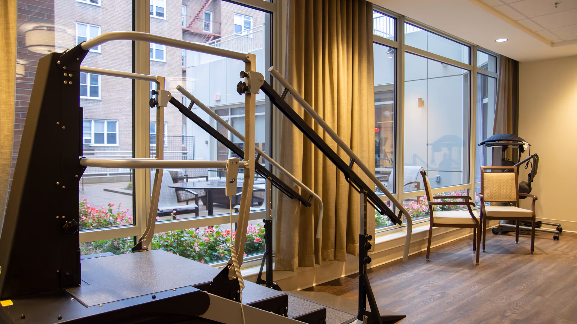 Rehabilitation gym area with parallel bars and chairs beside large windows.