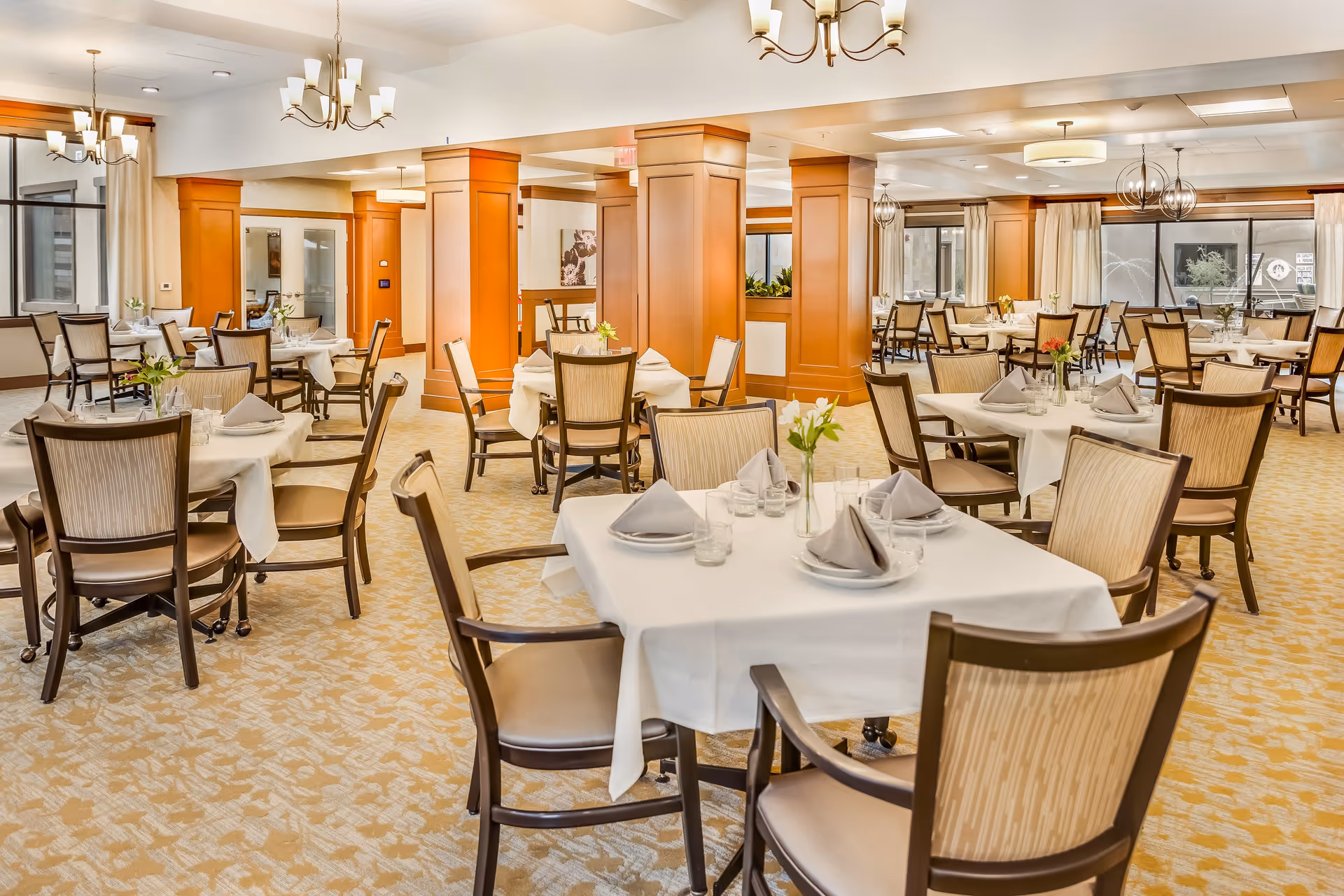 A spacious dining room in Legacy Village of Salt River with multiple tables covered in white tablecloths, each set with plates, glasses, and folded napkins. The room features beige patterned carpet, wooden chairs with cushioned seats, large windows with curtains, and warm lighting from chandeliers and ceiling fixtures.