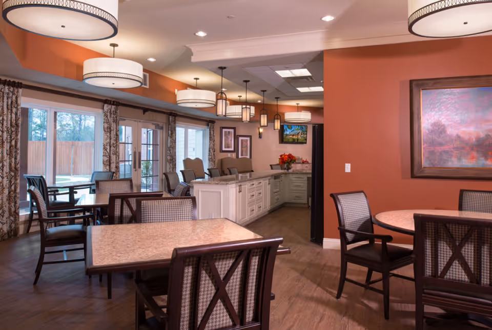 Communal dining room with multiple tables and chairs, pendant lights, large windows, and a serving counter in the background.