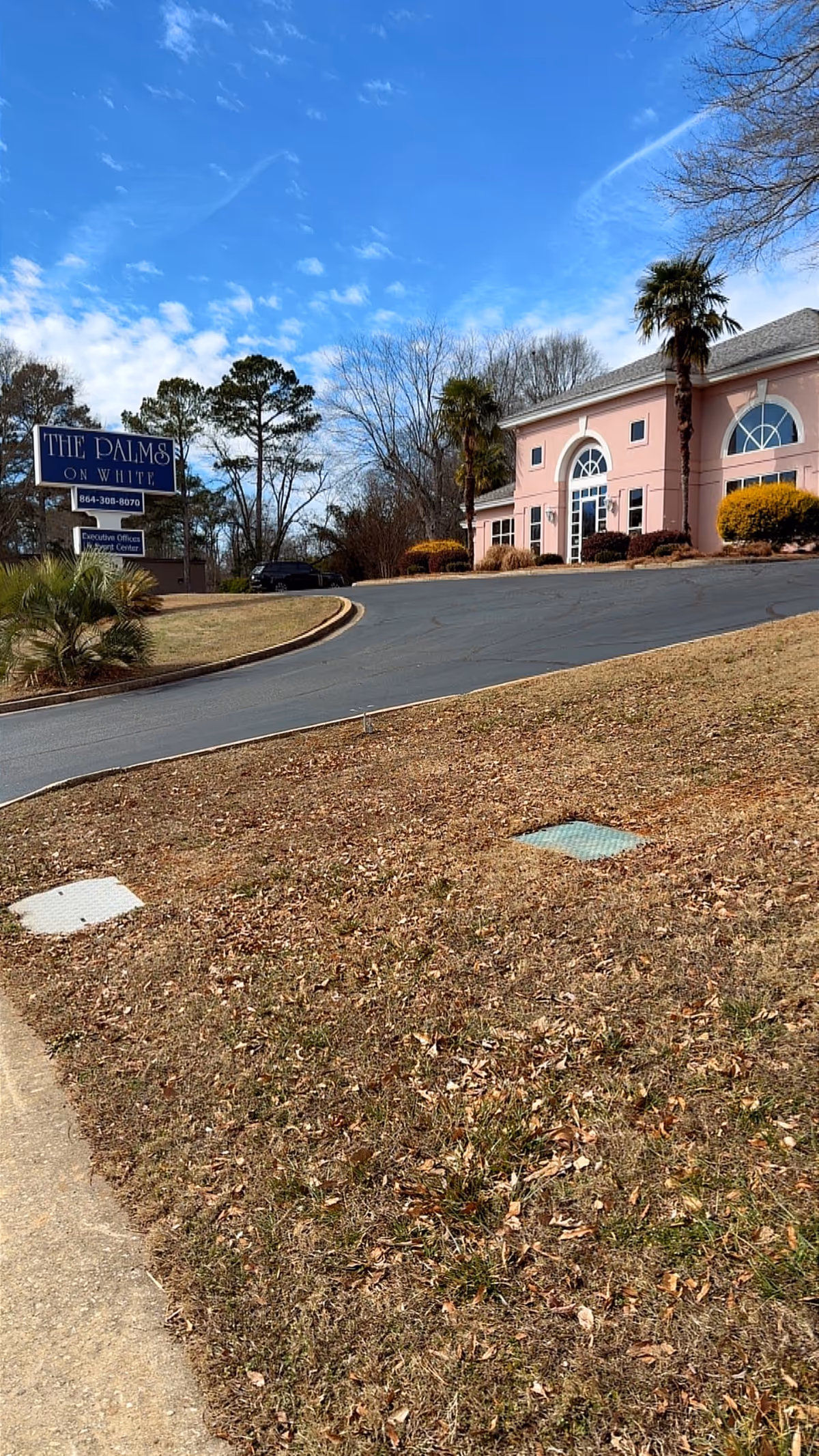 Pink senior living facility with arched windows, sign, palm trees, and curved driveway.