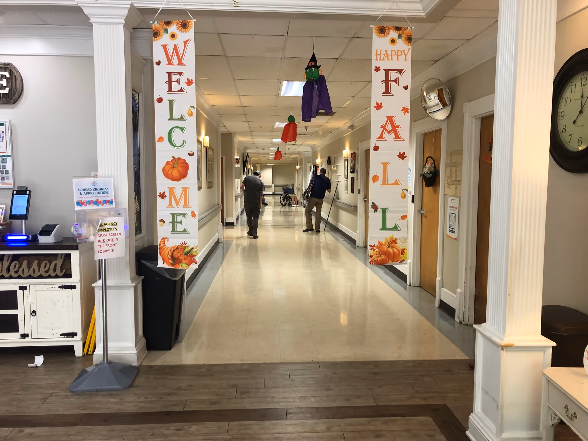 Wide hallway inside a senior care facility with Welcome Fall banners, clocks, and people walking.