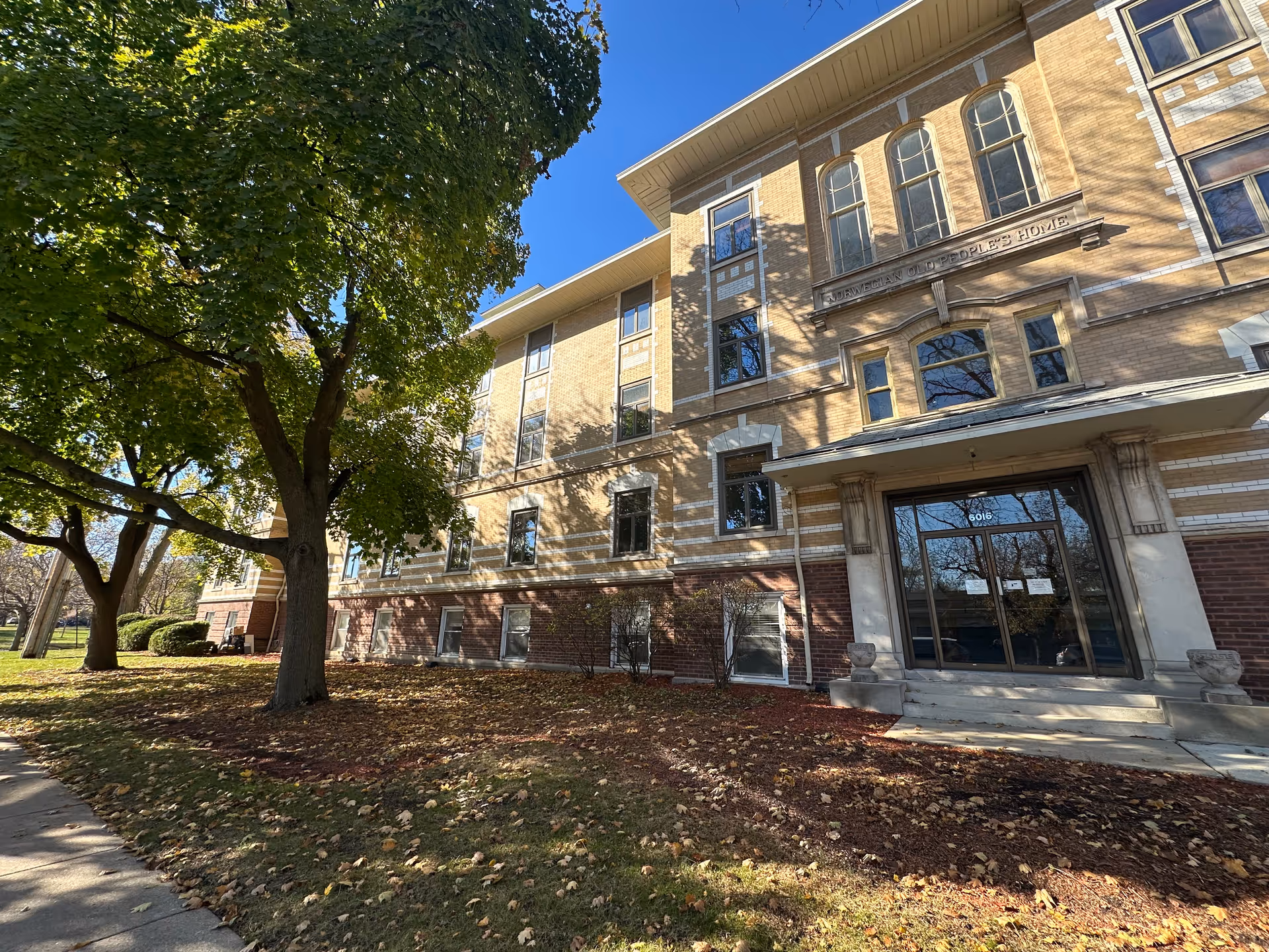 Front exterior of a multi-story brick senior living building with trees, a sidewalk, and a glass entrance.