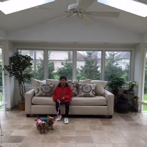Person sits on a beige sofa in a bright living room with plants and dogs.