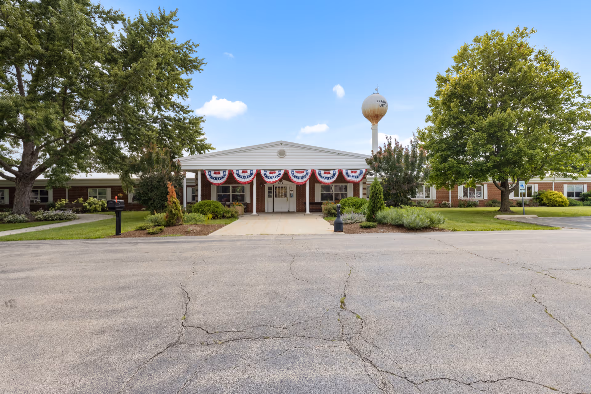 Front entrance of a single-story brick senior living facility decorated with patriotic bunting, flanked by trees and a water tower in the background.