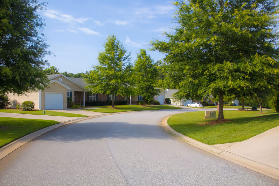 Curved residential street in a senior living community lined with single-story homes, green lawns, and trees.