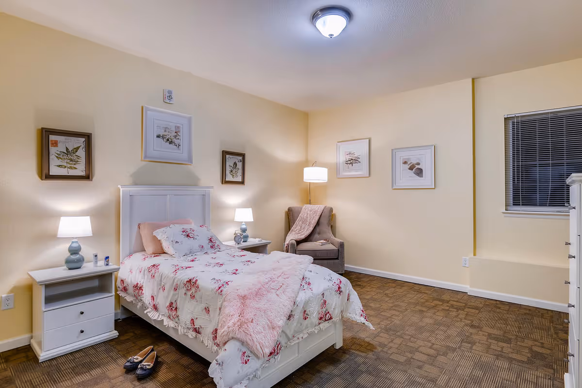 A cozy pastel bedroom with a bed, nightstands, lamps, framed art, and a chair.