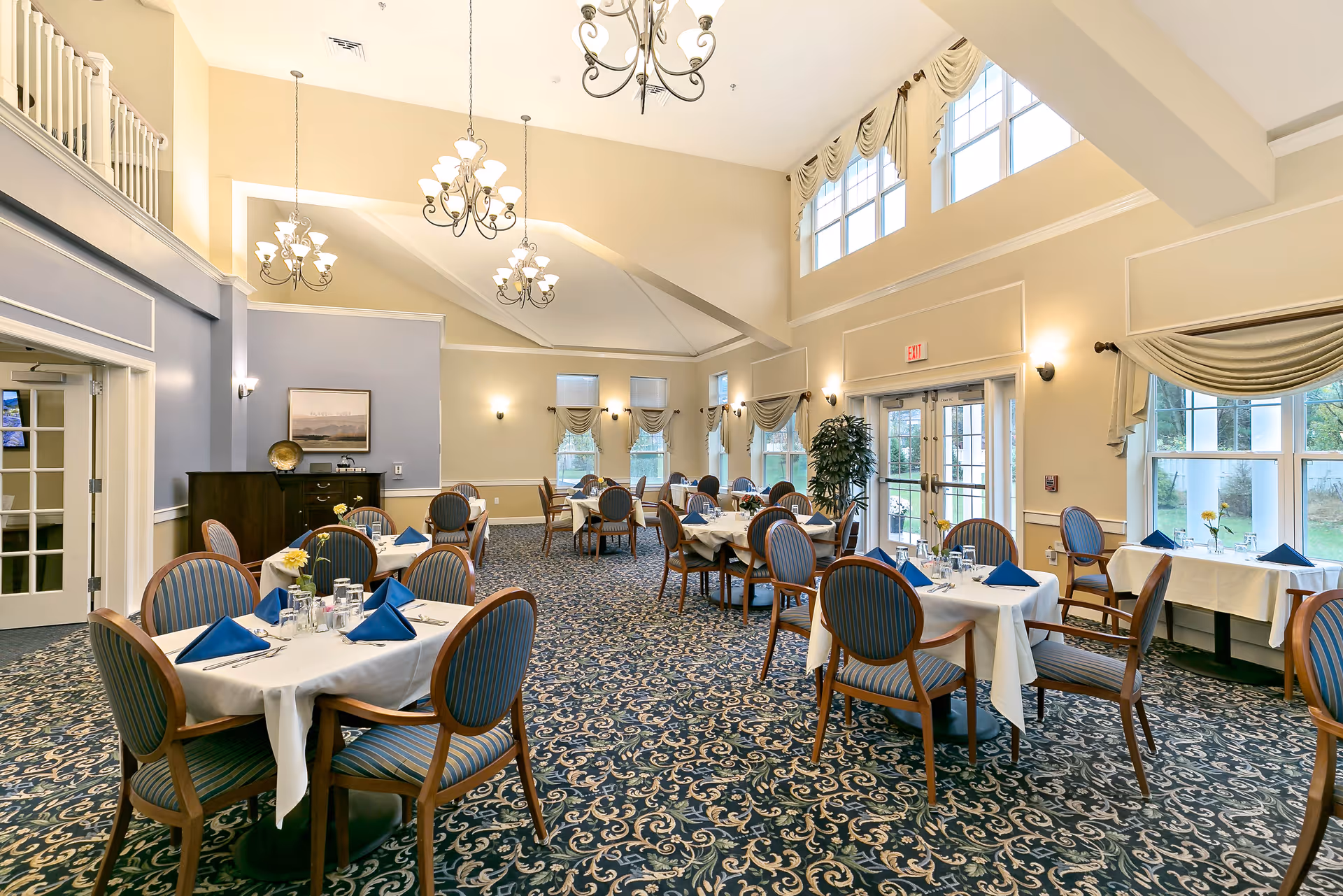 A spacious dining room with multiple round tables covered with white tablecloths and set with blue napkins, glasses, and silverware. The room features large windows with beige drapes, patterned carpet, and elegant chandeliers hanging from a high ceiling. There are wooden chairs with blue striped cushions around the tables, and a sideboard with decorative items against one wall.