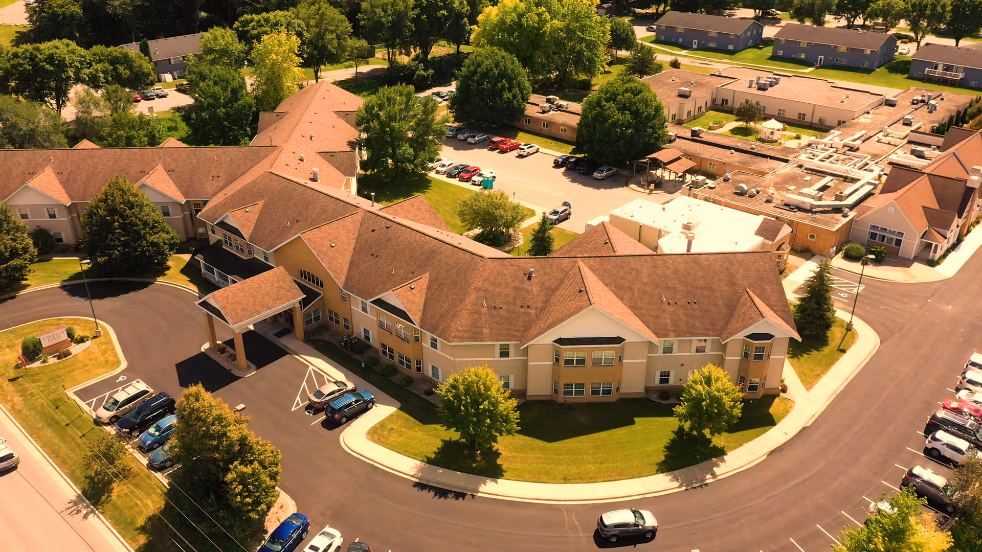 Aerial view of Laurels Edge Assisted Living facility showing a large, L-shaped building with a covered entrance, surrounded by green lawns, trees, and parking lots with several cars parked.