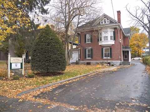 Brick senior living facility on a tree-lined street with a driveway and fallen autumn leaves.
