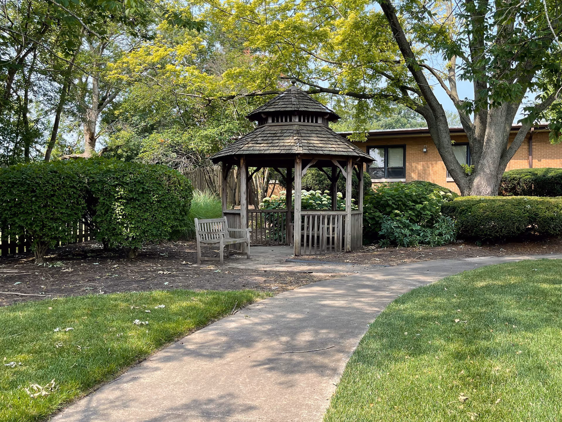 A wooden gazebo with a shingled roof situated in a garden area surrounded by green bushes and trees. A wooden bench is placed next to the gazebo. A curved concrete pathway leads to the gazebo, and a brick building is visible in the background.
