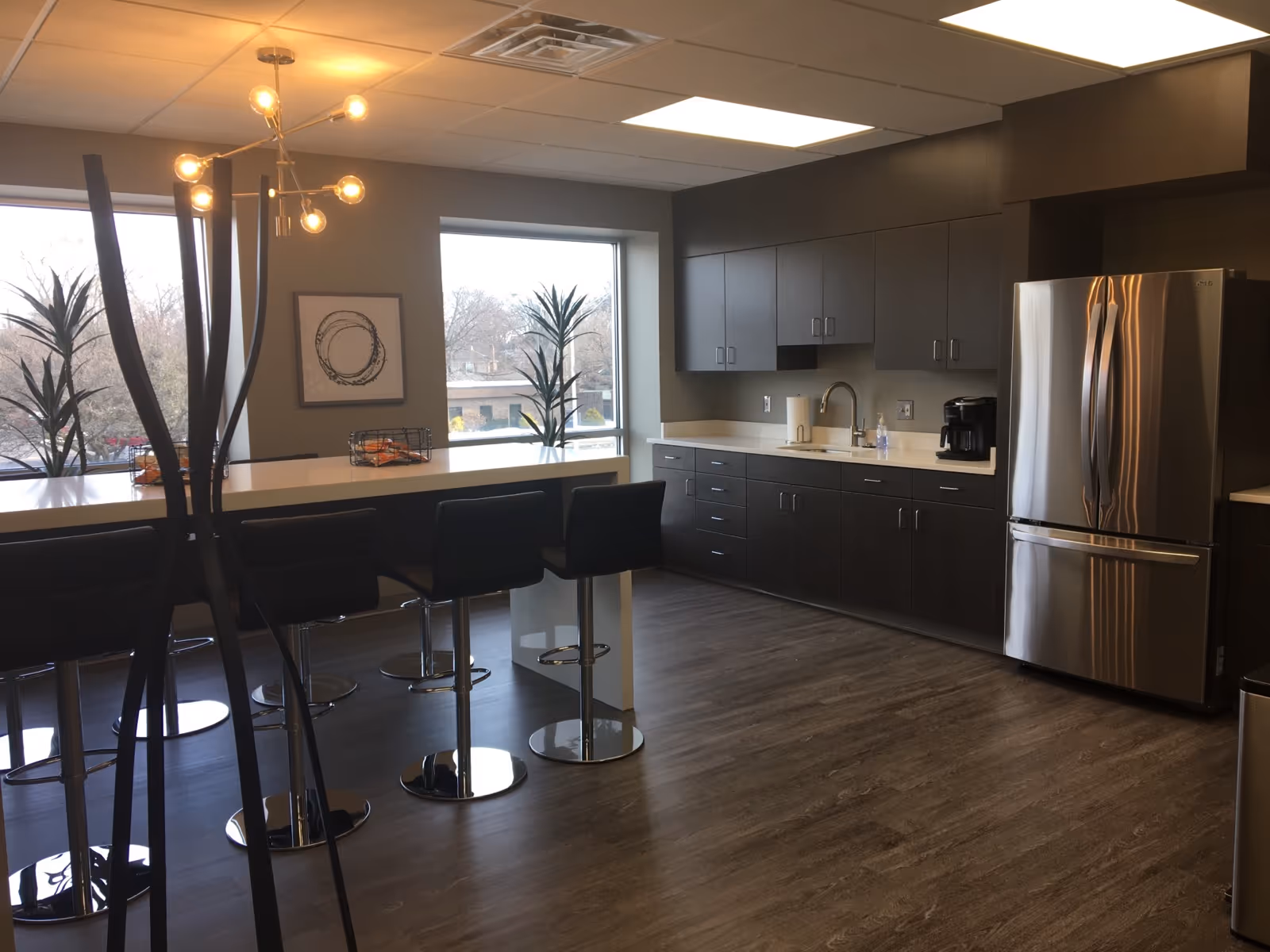 A modern facility kitchen with dark cabinets, stainless refrigerator, sink, and row of high stools.