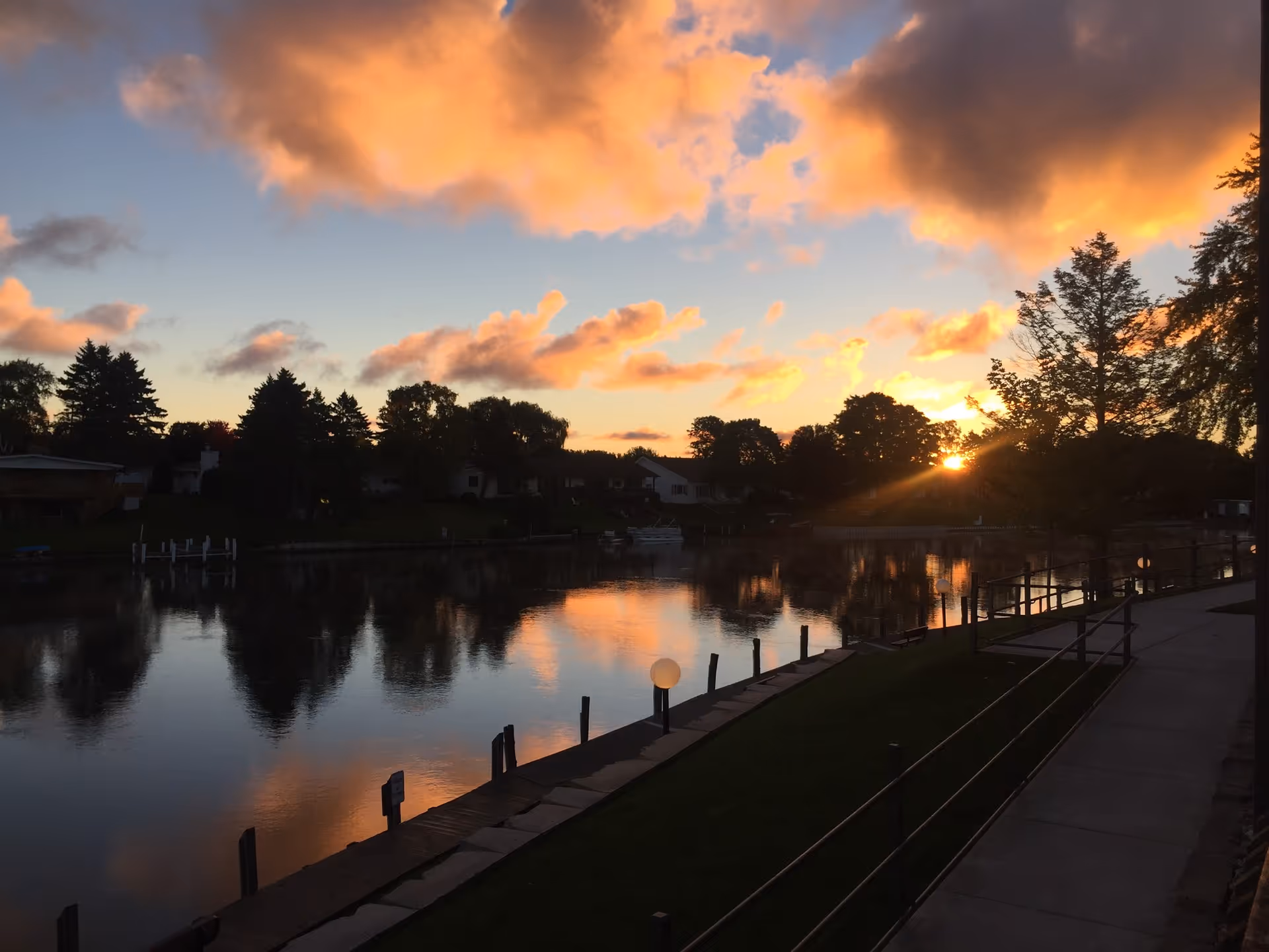 Sunset over a calm lake with trees, a walkway, and waterfront railing.
