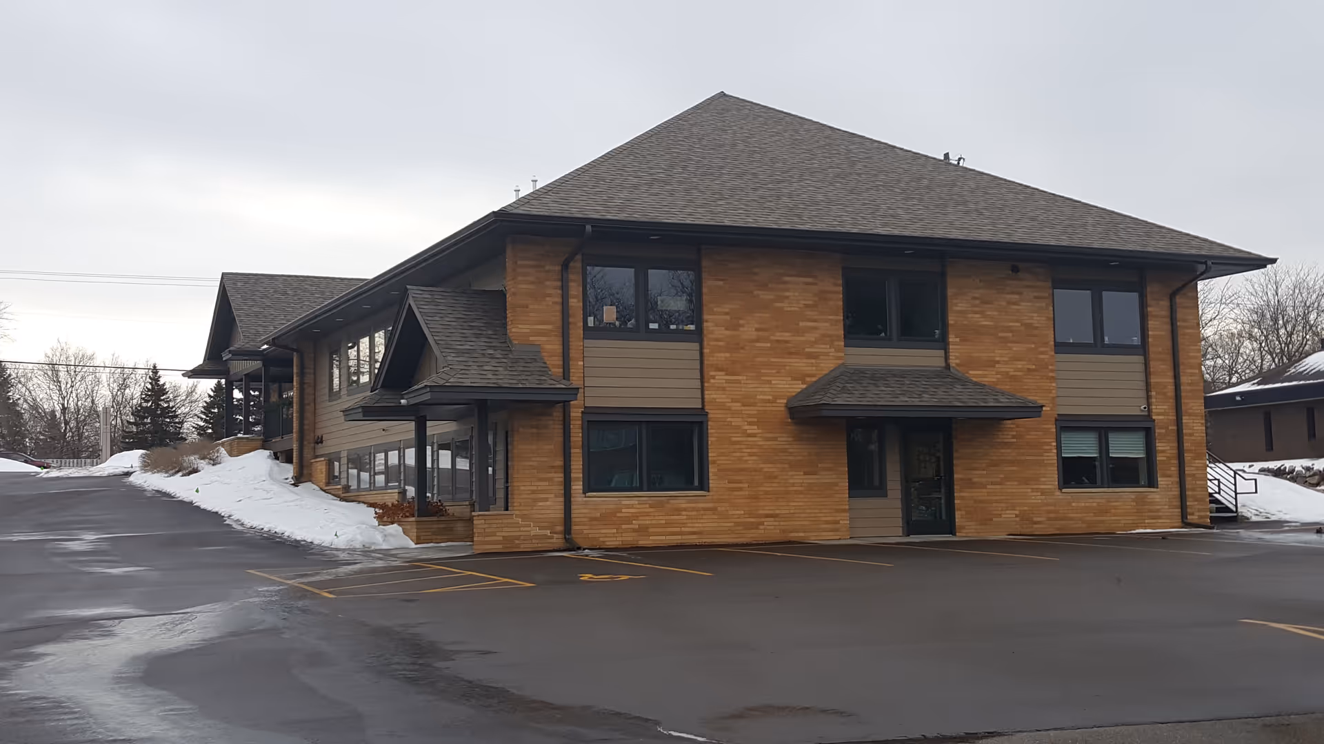 Two-story brick medical facility with a sloped roof, large windows, a paved parking lot, and snow.