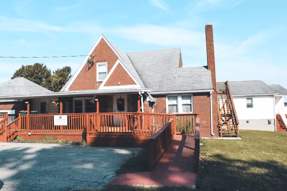 Exterior view of a red brick building with a gray shingled roof and a large wooden porch with railings and a wheelchair ramp. The building has a chimney and an external staircase on the right side. There is a sign on the porch that reads 'Countryside Personal Care Home'. The sky is clear and blue with some scattered clouds.