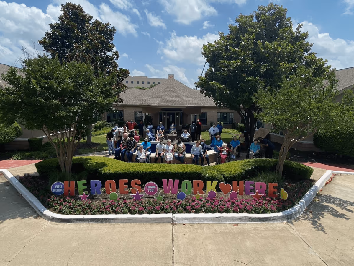 A group of residents and staff gather in the front courtyard of a senior living building behind a flowerbed with colorful 'HEROES WORK HERE' signs.