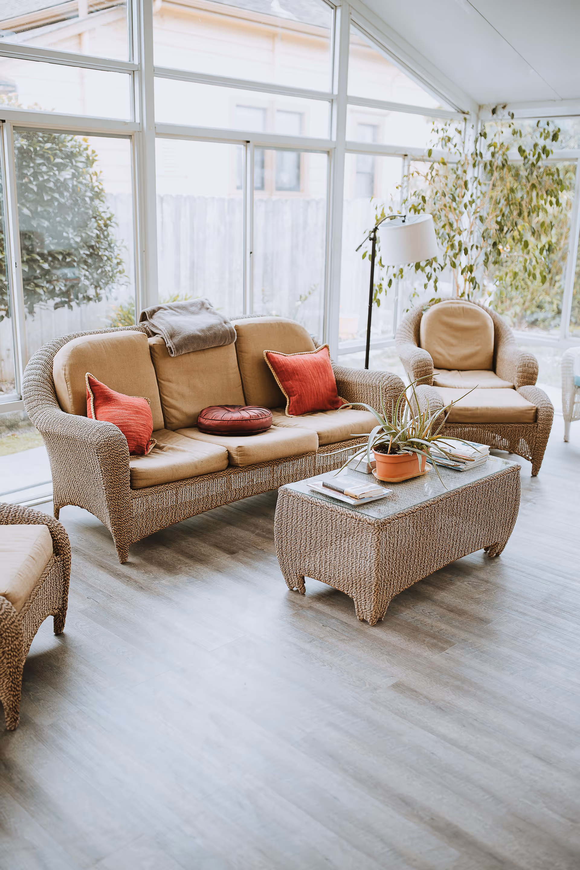 A bright and cozy living room area with wicker furniture including a sofa, armchair, and coffee table. The sofa has beige cushions and red throw pillows, with a folded blanket on the backrest. The coffee table holds a potted plant and some magazines. Large windows let in natural light and show greenery outside.