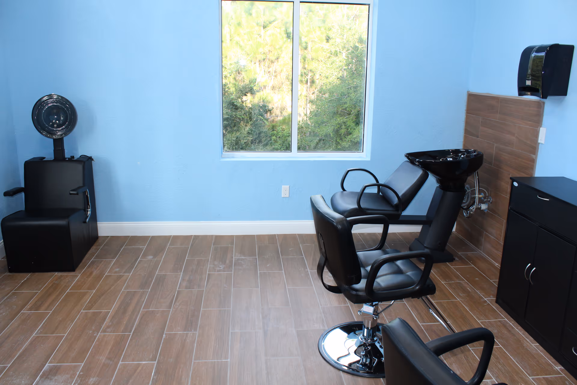 Blue-walled senior living salon with black barber chairs, a washing station, and window view.
