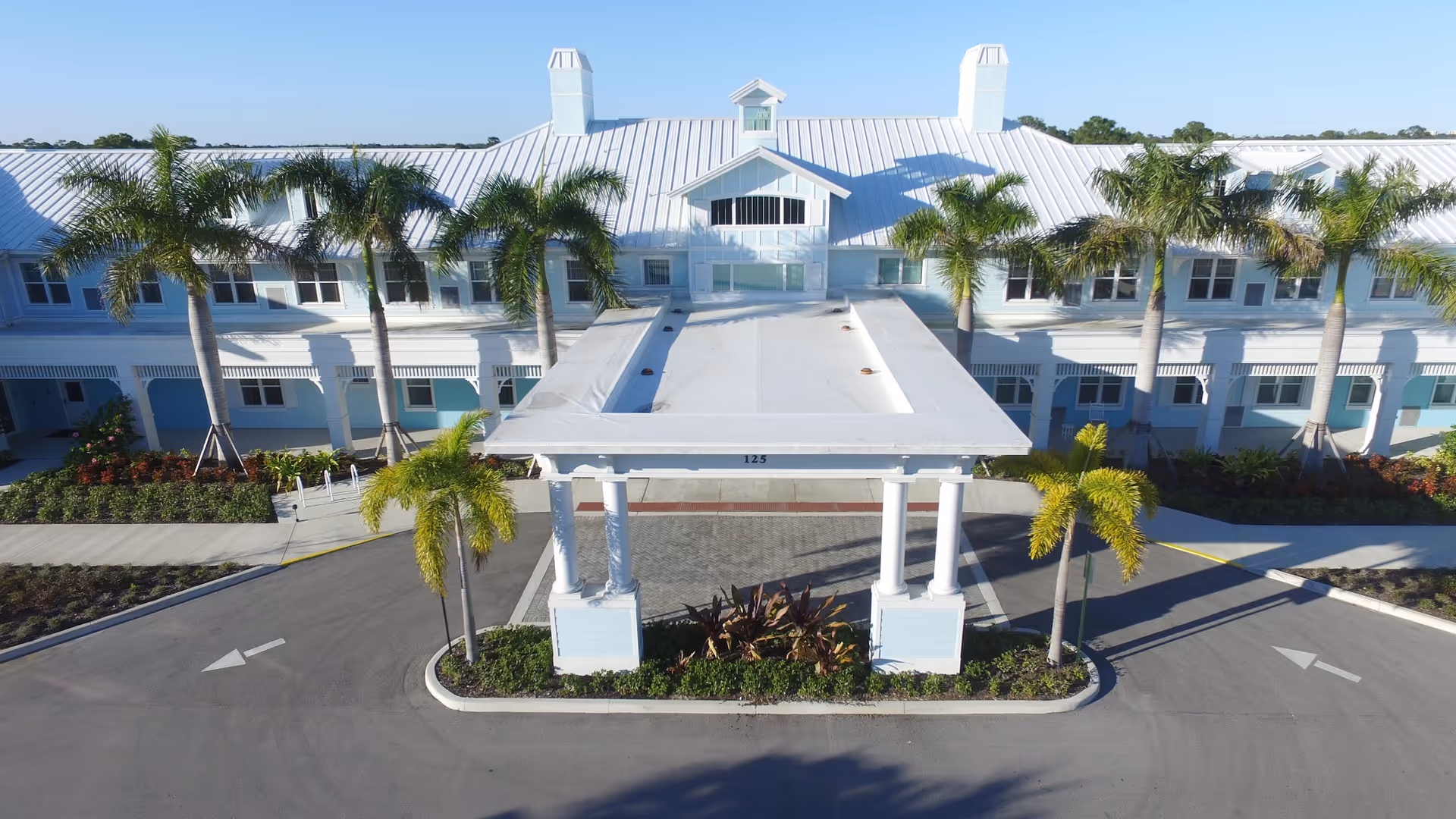 Front exterior view of a large white building with a metal roof, palm trees lining the entrance, and a covered drop-off area with columns in front.