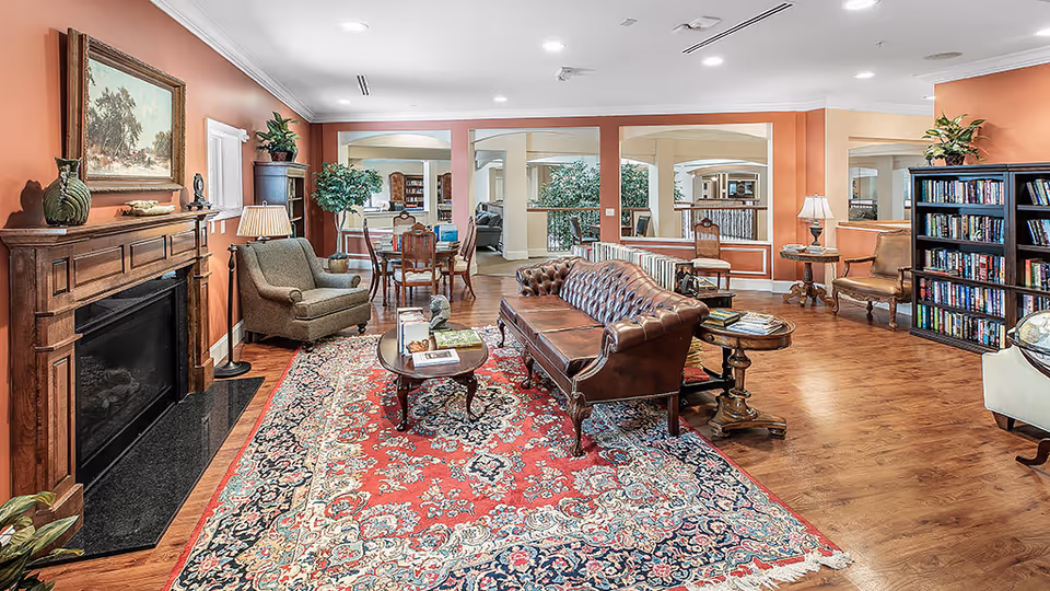 A cozy senior living facility common area with a brown leather sofa, an armchair, wooden coffee tables, a large patterned rug, a wooden fireplace with decorative items, bookshelves filled with books, and warm orange walls. The room has wooden flooring and is well-lit with ceiling lights and table lamps.