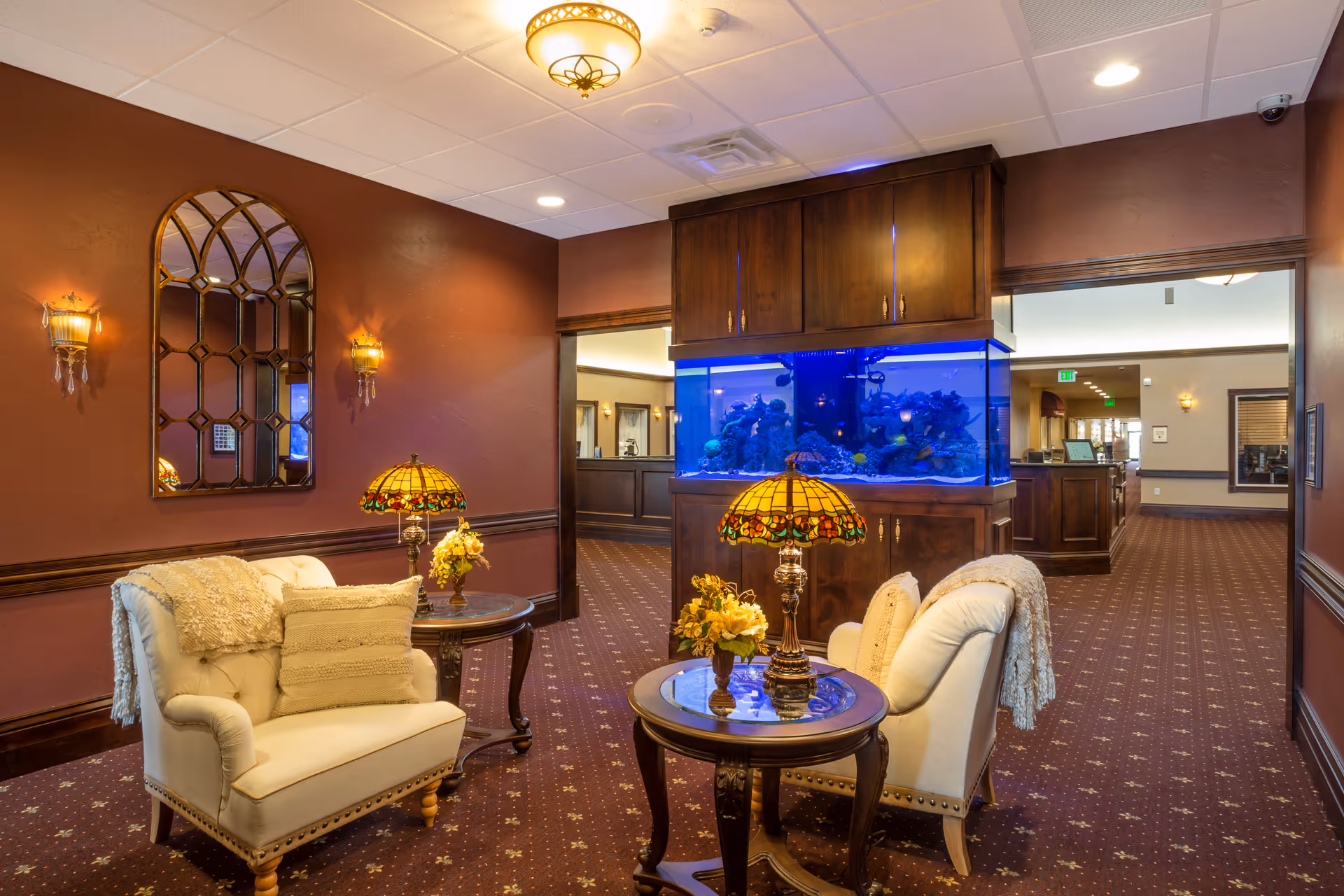 A cozy lounge area with upholstered chairs, side tables topped with Tiffany lamps, and a large built-in aquarium in a carpeted reception space.