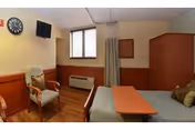 Small rehabilitation room with a single bed and overbed table, armchair, wall-mounted TV, window, privacy curtain, and wooden cabinetry.