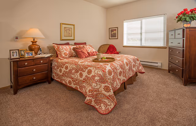A cozy bedroom with a bed covered in a red and beige patterned quilt and multiple pillows. There is a wooden nightstand with a lamp and framed photos on the left side of the bed. On the right side of the room, there is a wooden dresser with a vase of red flowers on top. The room has beige carpet and walls, two framed pictures on the walls, and a window with blinds letting in natural light.