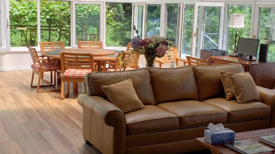 Bright sunlit living area with a beige sofa, coffee table, and wooden dining set.
