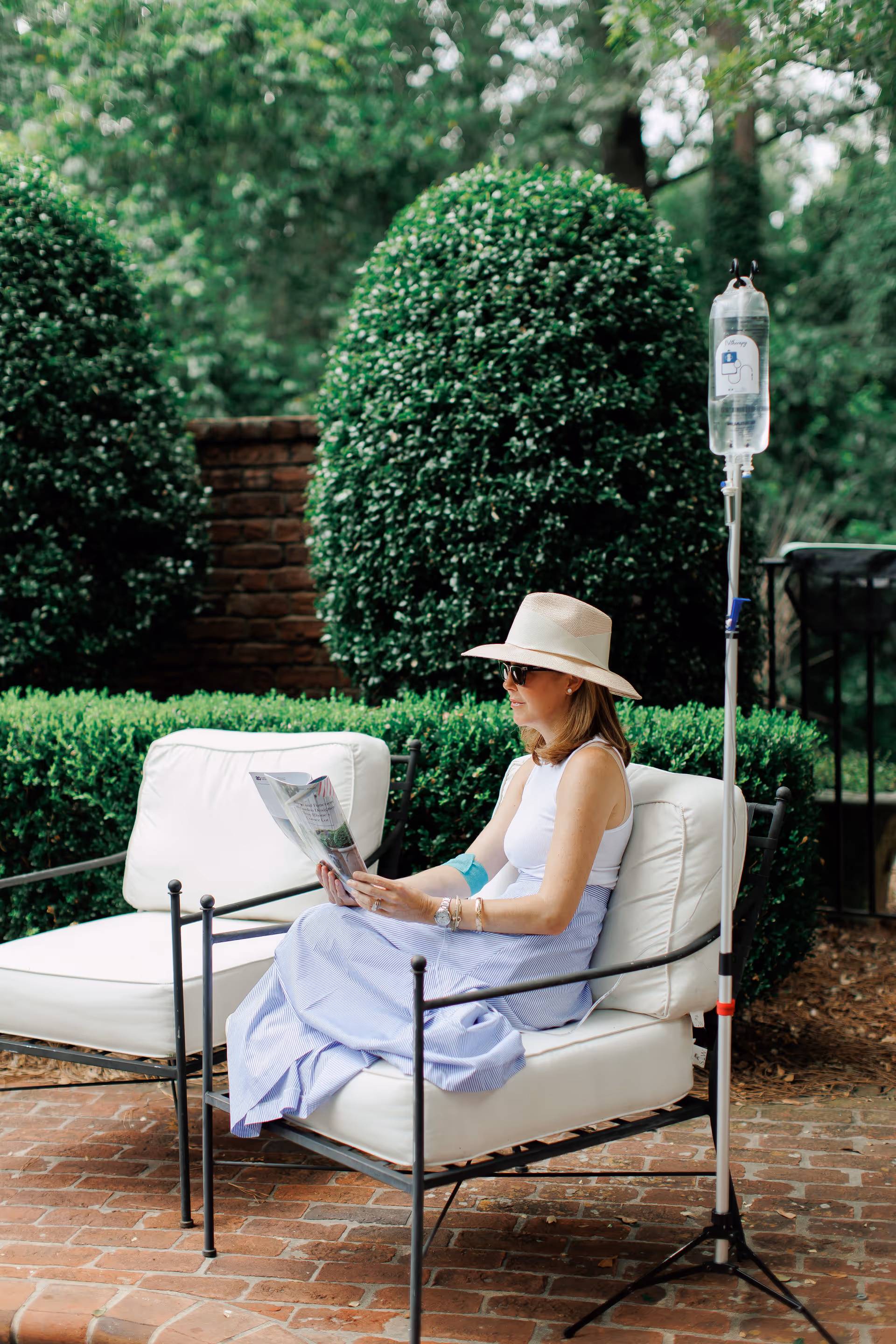 A woman wearing a hat sits outdoors reading a magazine beside an IV stand.