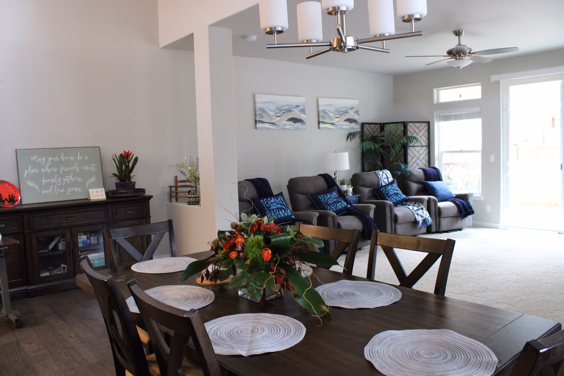 A bright and spacious living and dining area with a dark wooden dining table set with six chairs and round placemats. A floral centerpiece decorates the table. In the background, there are three gray recliner chairs with blue pillows and throws, a floor lamp, and two landscape paintings on the wall. Large windows and a glass door let in natural light, and a ceiling fan and modern light fixture hang from the ceiling. A wooden cabinet with plants and decorative items is visible on the left side.