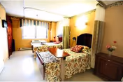 Shared patient room with two floral beds, nightstands, a dresser, and a window at the end.
