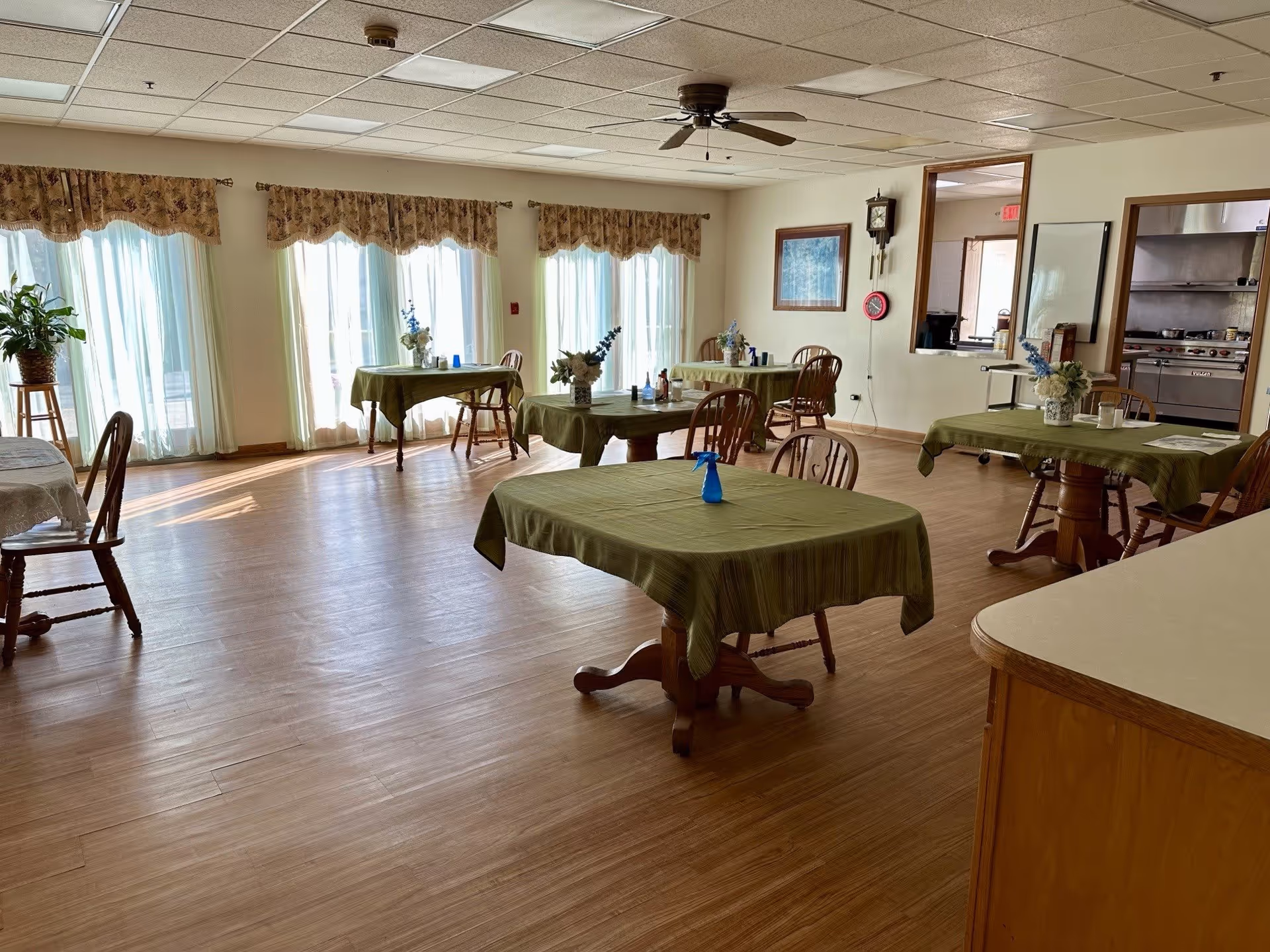 A dining room with several wooden tables covered with green tablecloths and wooden chairs around them. The room has large windows with sheer curtains and floral valances, allowing natural light to fill the space. There are flower arrangements on the tables and a ceiling fan overhead. A kitchen area is visible through an open window in the wall.