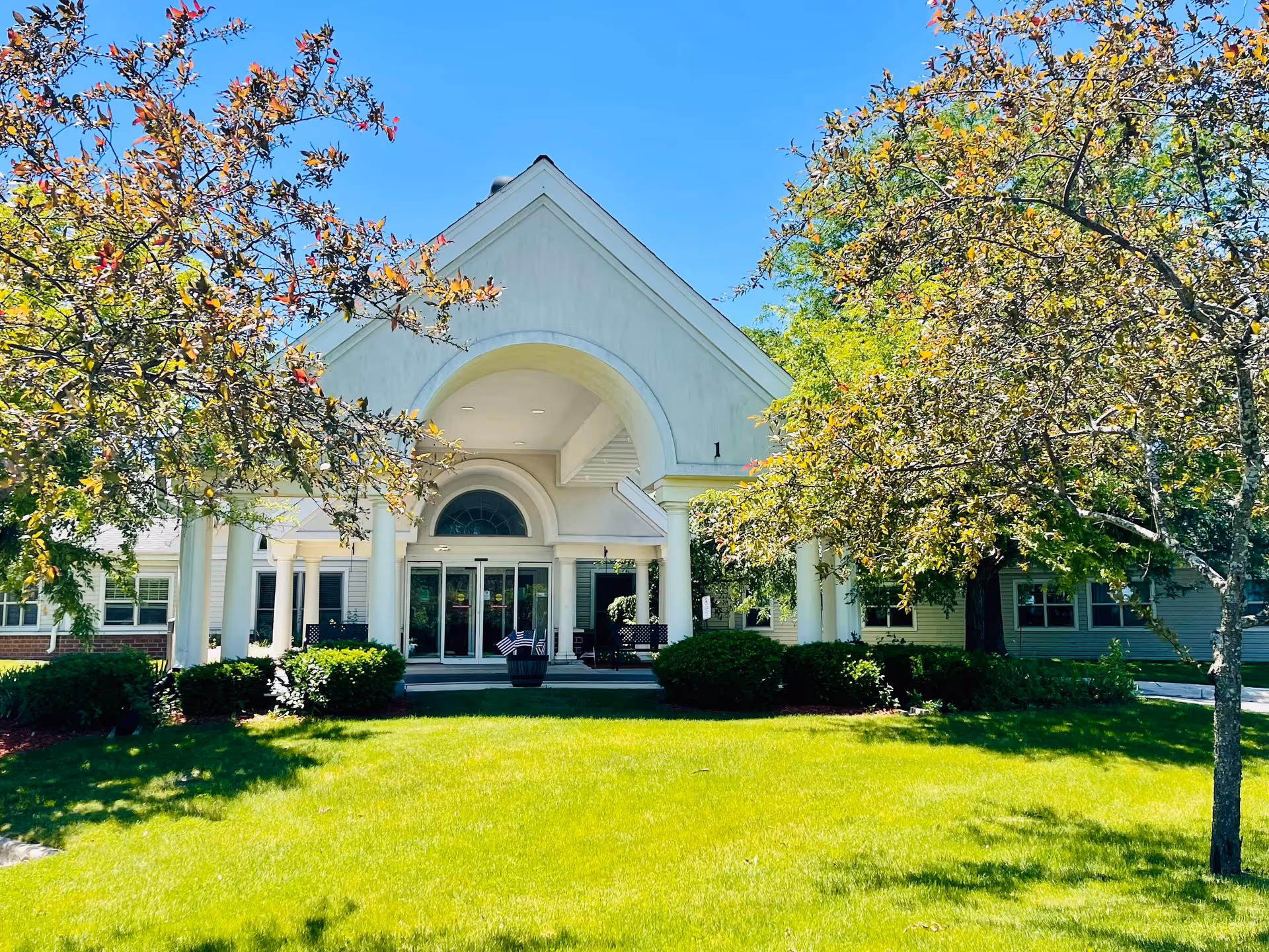 Front entrance of a white care and rehab building with columns, glass doors, and a green lawn framed by trees.