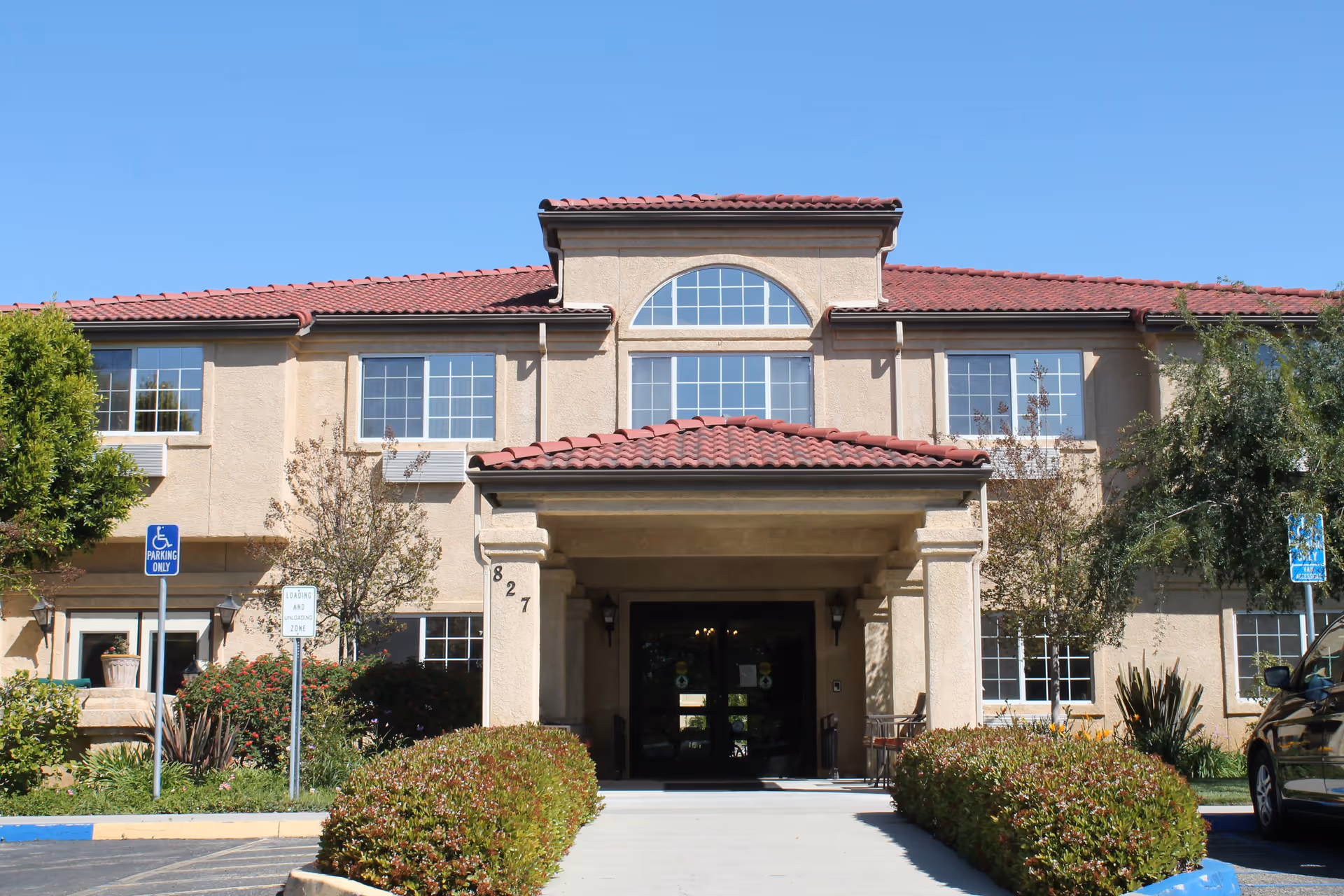 Front exterior view of a two-story building with beige stucco walls and a red tile roof under a clear blue sky. The entrance has a covered porch with columns and the number 827 displayed on one column. There are bushes and small trees on either side of the walkway leading to the entrance, and a parking lot with handicap parking signs is visible.